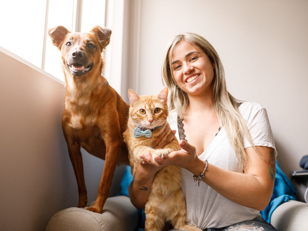 Mulher sorrindo com cachorro e gato em ambiente doméstico, representando bem-estar, convivência e saúde mental pet.